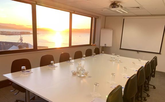 A conference room set up with a big table, six rolling desk chairs along opposite sides, pens, note pads, water glasses and pitchers.