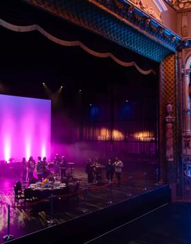 The stage at St James Theatre in Wellington. People are standing on stage and a few tables and chairs are set up for an event. The house lights are off and the room is illuminated with purple and blue spotlights.