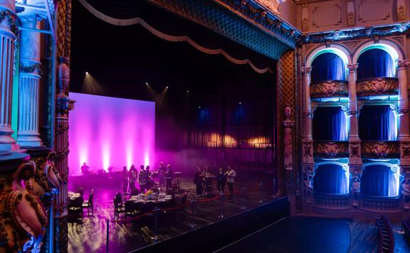 The stage at St James Theatre in Wellington. People are standing on stage and a few tables and chairs are set up for an event. The house lights are off and the room is illuminated with purple and blue spotlights.