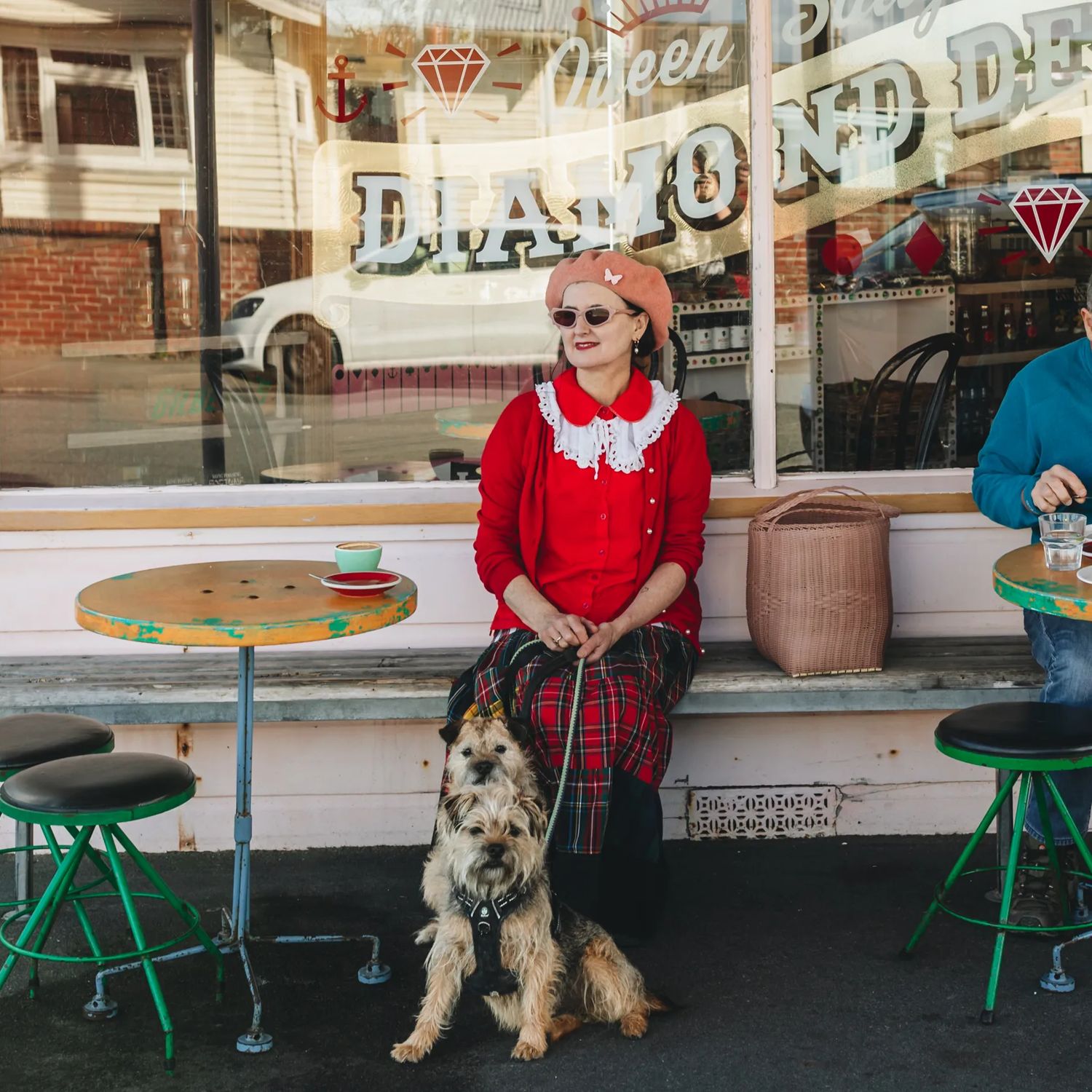 Person wearing a red outfit sitting outside Queen Sally's Diamond Deli cafe, holding the lead attached to two small tan coloured dogs.