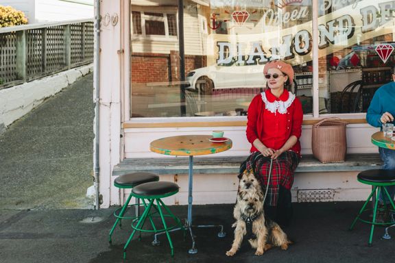 Person wearing a red outfit sitting outside Queen Sally's Diamond Deli cafe, holding the lead attached to two small tan coloured dogs.