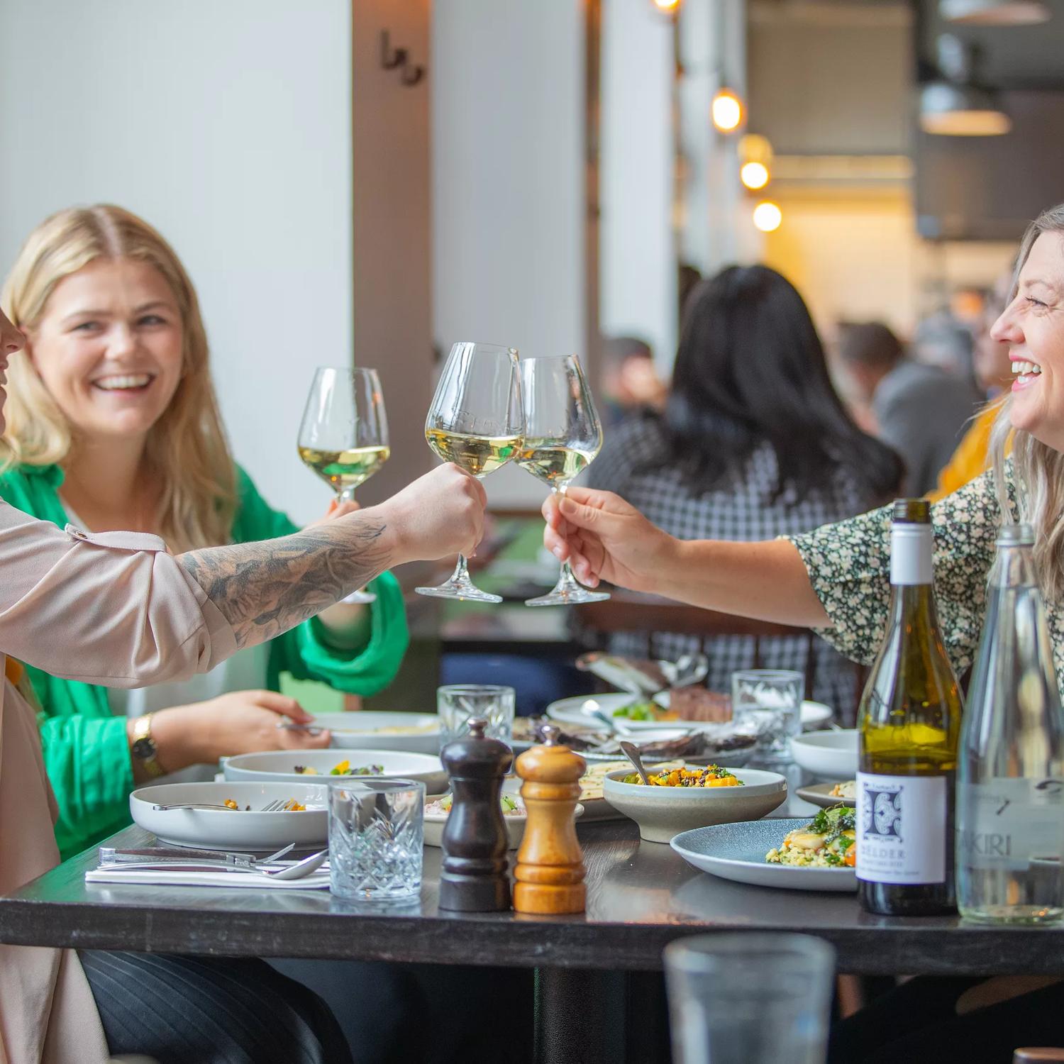 Three people sit around a table covered in foot plates at Charley Noble. They are clinking wine glasses.