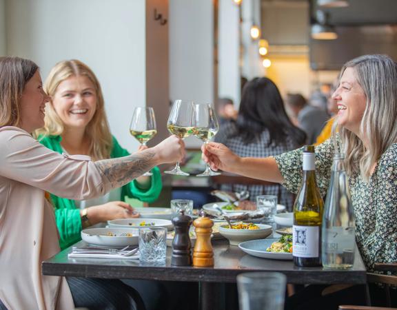 Three people sit around a table covered in foot plates at Charley Noble. They are clinking wine glasses.