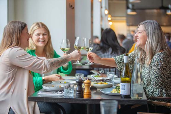 Three people sit around a table covered in foot plates at Charley Noble. They are clinking wine glasses.