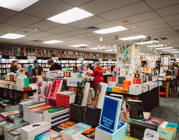 Shoppers inside Unity Books, a bookstore in Wellington. The large book-filled space has grey carpets, fluorescent lights and a white panelled ceiling. 