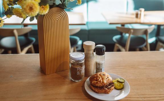 A cheese scone and a couple of pickle slices on a small white plate with three seasoning shakers and a vase with yellow flowers on a table at Pickle & Pie.