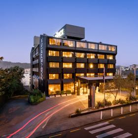 An exterior view of the Mövenpick Hotel at dusk, looking from The Terrace towards the black building. The windows are lit up and a blur of car lights snakes up the driveway.