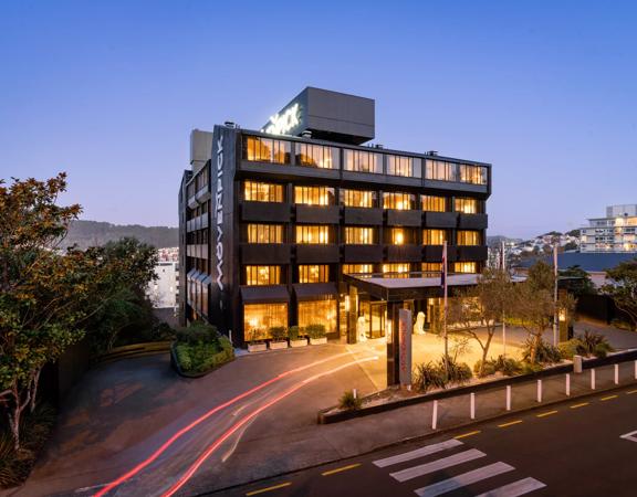 An exterior view of the Mövenpick Hotel at dusk, looking from The Terrace towards the black building. The windows are lit up and a blur of car lights snakes up the driveway.