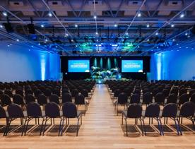 Hundreds of chairs face toward a stage with small trees on it, blue LED lights full the room.