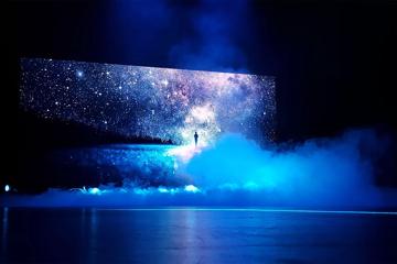 A starry night sky and a person’s silhouette are projected onto a large screen with smoke billowing I the foreground.