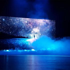 A starry night sky and a person's silhouette are projected onto a large screen with smoke billowing I the foreground. 
