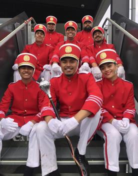 Eight marching band members from the film Red White and Brass are uniformed in red jackets, red hats and white pants sit together on stairs smiling.