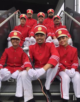 Eight marching band members from the film Red White and Brass are uniformed in red jackets, red hats and white pants sit together on stairs smiling. 