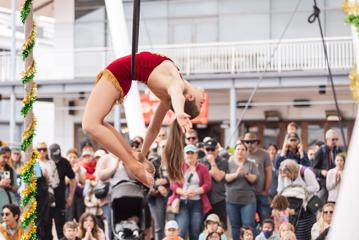 An acrobat in a red leotard performs a trick suspended in mid-air in front of an applauding audience.