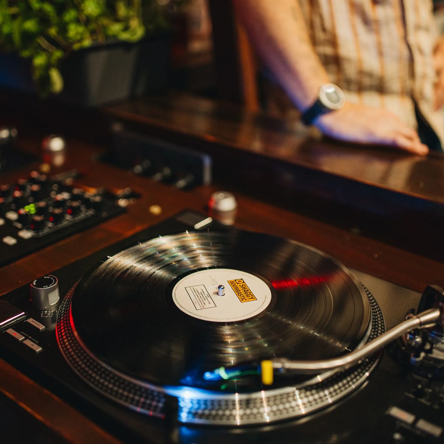 Close-up of a vinyl record spinning on a turntable, with a mixer and controls nearby and a person leaning on the counter in the background under warm lighting.