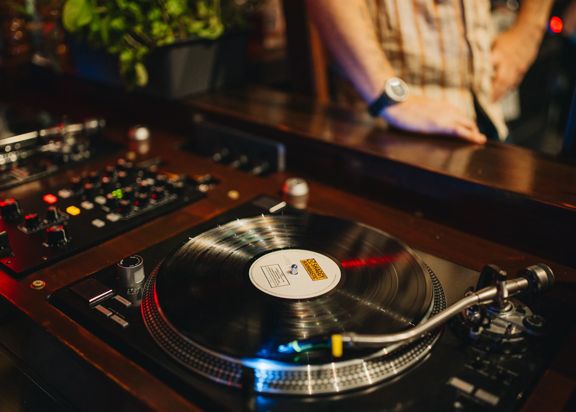Close-up of a vinyl record spinning on a turntable, with a mixer and controls nearby and a person leaning on the counter in the background under warm lighting.