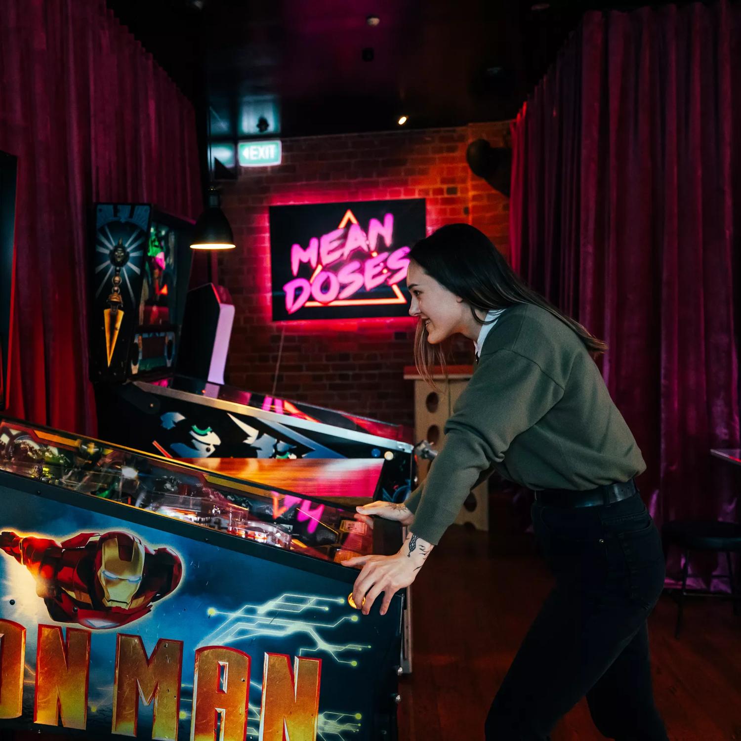 A person wearing black pants and a grey-green jumper is playing a Iron Man Pinball machine at Mean Doses Taproom, a bar in Te Aro, Wellington. The walls are covered by merlot-coloured velvet curtains and the dark space is illuminated by neon pink light.