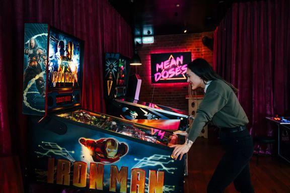 A person wearing black pants and a grey-green jumper is playing a Iron Man Pinball machine at Mean Doses Taproom, a bar in Te Aro, Wellington. The walls are covered by merlot-coloured velvet curtains and the dark space is illuminated by neon pink light.