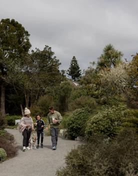 Two adults and a child walking on path with dog on lead. Walking through a garden with mature trees and plants, wearing winter clothes. Grey sky, some houses in the background.