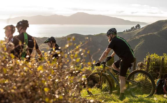 Four mountain bikers at the top of the Red Tape Track on Whareroa Farm, overlooking Kāpiti Coast and Kapiti Island.