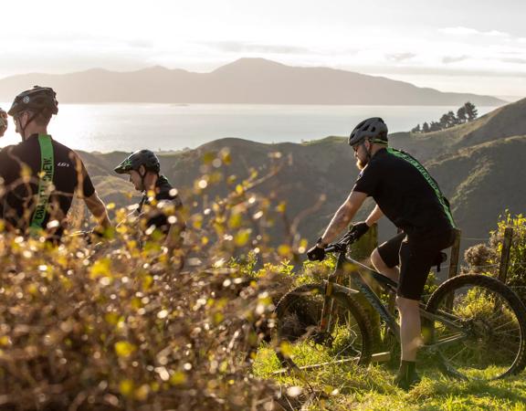 Four mountain bikers at the top of the Red Tape Track on Whareroa Farm, overlooking Kāpiti Coast and Kapiti Island.