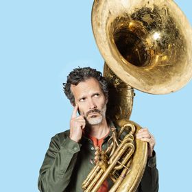 Bret McKenzie holds a tuba with a pensive expression.