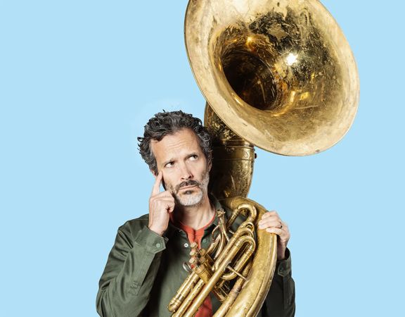Bret McKenzie holds a tuba with a pensive expression.