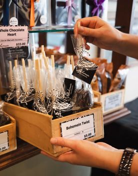 A wooden box of Bohemein Twists, a chocolate treat on a stick, on the shelf at Bohemein Fresh Chocolates. A person is picking one Bohemein Twist with their right hand and holding the box with their left.