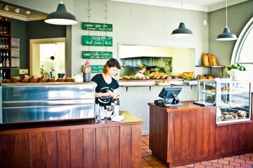 A café with pale green walls and a wooden counter. A barista wearing black shirt is making a coffee.