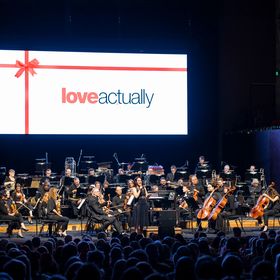 A live orchestra on stage with the title card of the film Love Actually projected on a screen above them.