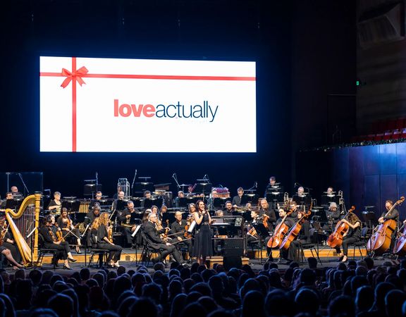 A live orchestra on stage with the title card of the film Love Actually projected on a screen above them.