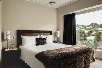 A room at the Bolton Hotel in Wellington with a queen-sized bed, brown fur throw blanket, two bedside tables with a lamp on each and a large window with a view of houses on a hillside.