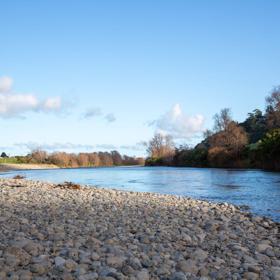 The Taitā Rock swimming hole in Lower Hutt, with lush green bush surrounding a blue river and large pebbles on the shore.