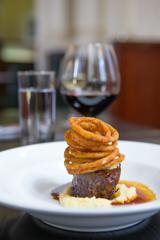 A filet mignon on a small bed of mashed potatoes and sauce with a stack of onion rings on top.