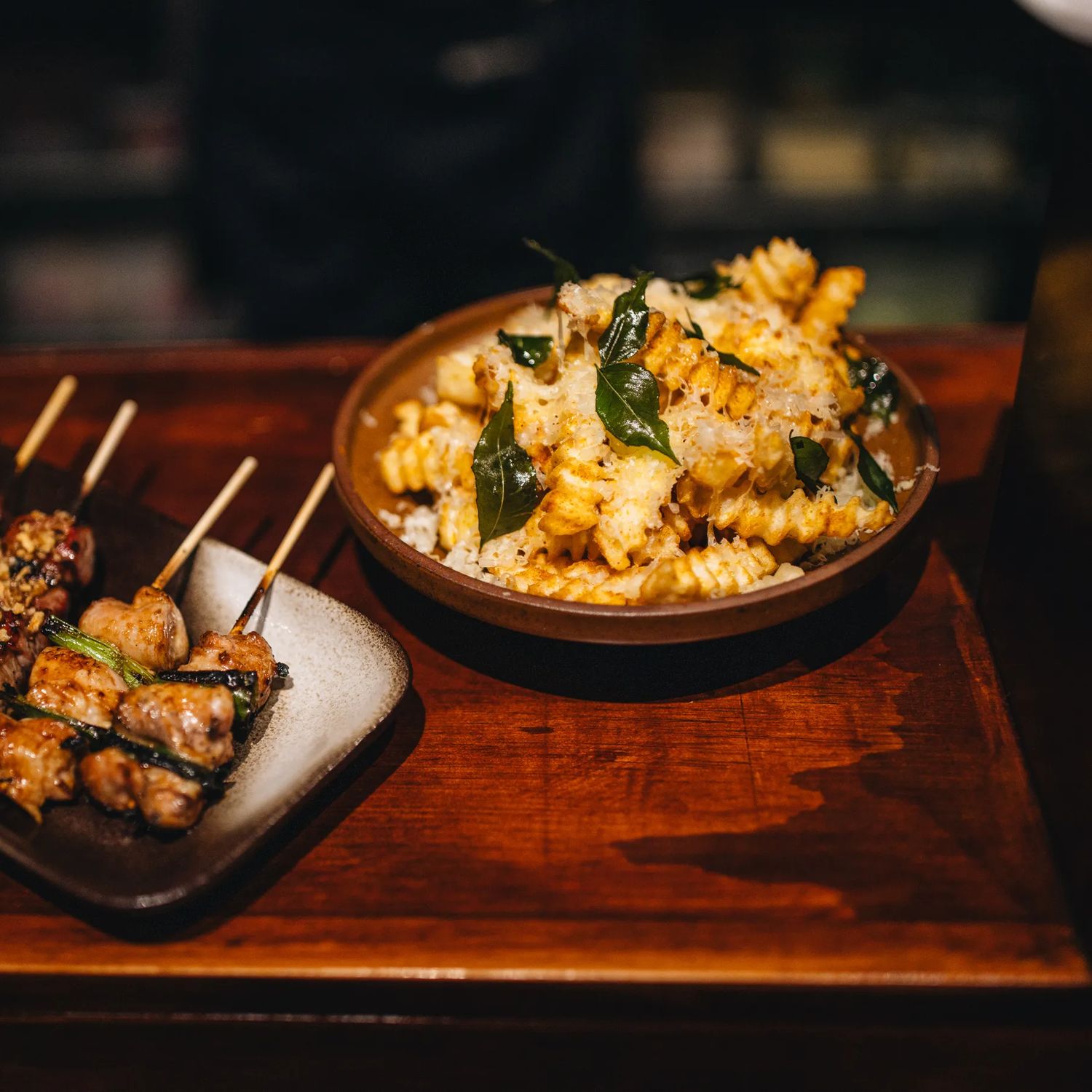 Two dishes sit on a wooden bar: skewers of grilled meat topped with herbs on a rectangular plate, and a bowl of pasta or rice mixed with vegetables and garnishes, both lit by warm, low lighting.