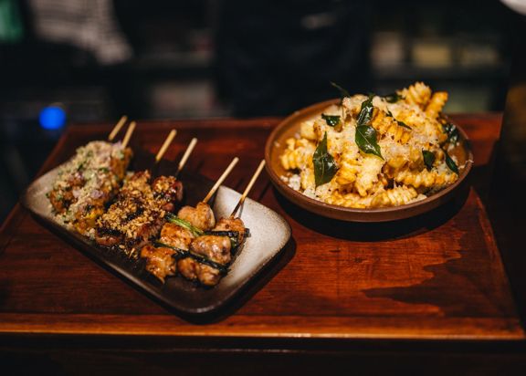Two dishes sit on a wooden bar: skewers of grilled meat topped with herbs on a rectangular plate, and a bowl of pasta or rice mixed with vegetables and garnishes, both lit by warm, low lighting.