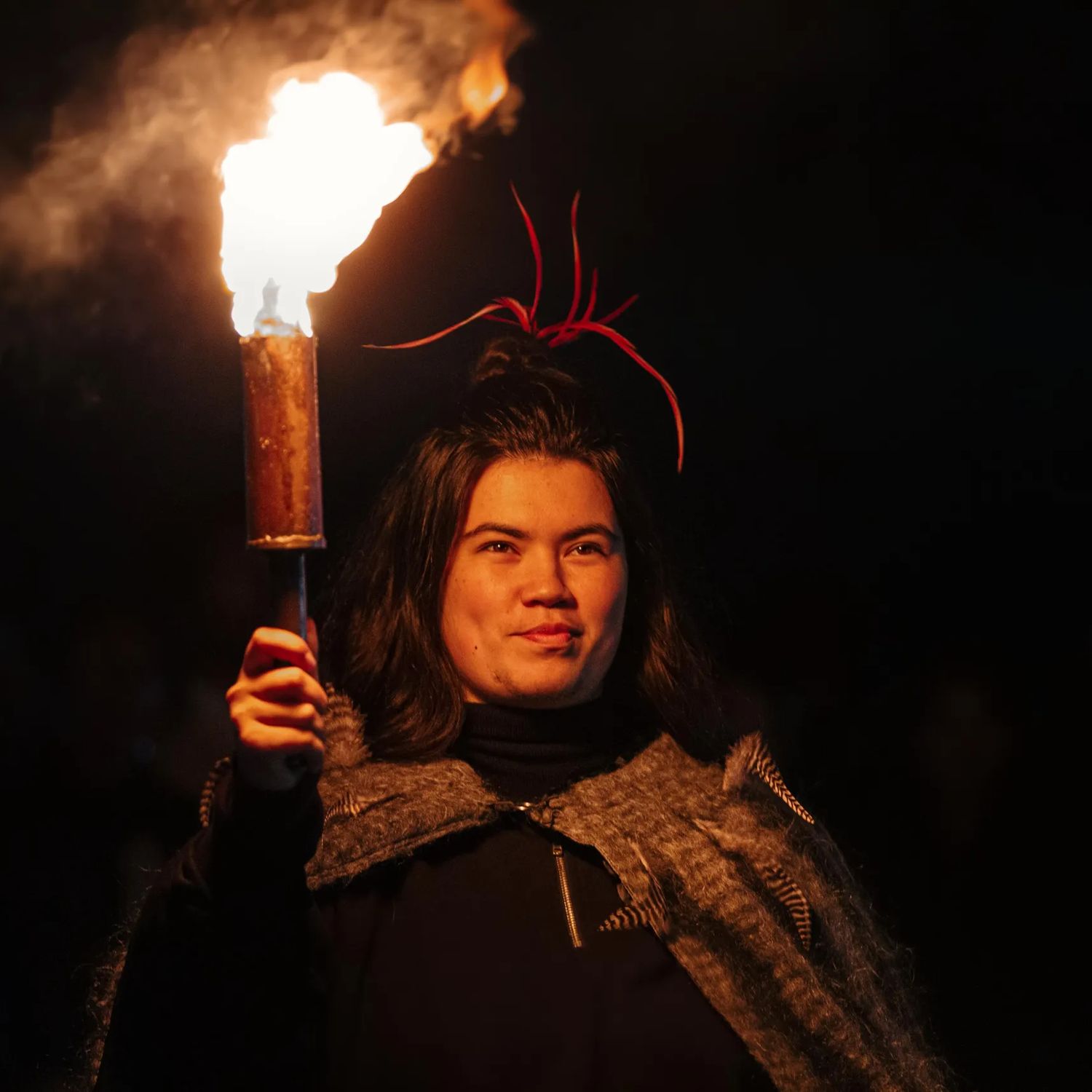 Cloaked in brown feathers, a Māori figure holds a glowing torch under the cover of night.