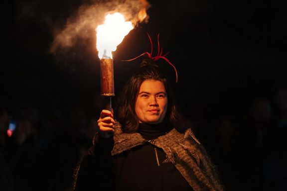 Cloaked in brown feathers, a Māori figure holds a glowing torch under the cover of night.