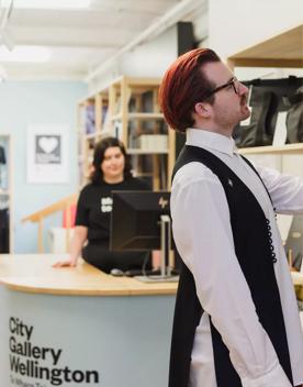 A red-haired man in a white collared shirt and black vest stands and touches a black leather backpack in the City Gallery Wellington giftshop. Behind him, a woman with brown hair wearing a black top stands behind a counter looking at a computer screen and
