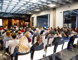 Inside the Te Ahumairangi Foyer of Tiakiwai Conference Centre, National Library of New Zealand. Around 50 people sit in chairs facing toward the speaker.