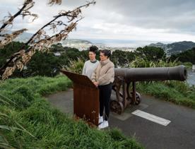 Two people stand by a cannon reading an information board on the Southern Walkway in Mount Victoria, Wellington. It is a grey, chilly day, the sky is cloudy and the city is visible in the distance.