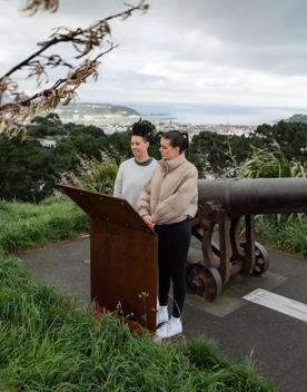 Two people stand by a cannon reading an information board on the Southern Walkway in Mount Victoria, Wellington. It is a grey, chilly day, the sky is cloudy and the city is visible in the distance.