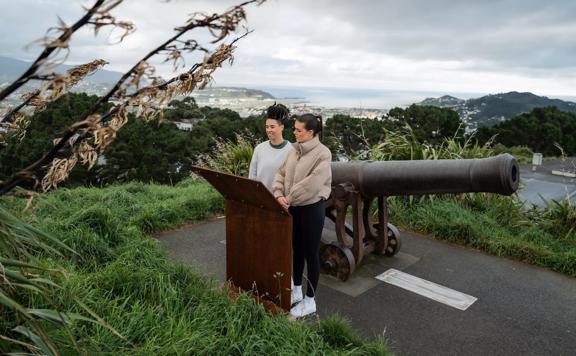 Two people stand by a cannon reading an information board on the Southern Walkway in Mount Victoria, Wellington. It is a grey, chilly day, the sky is cloudy and the city is visible in the distance. 