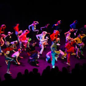 A group of 25 dancers wearing colourful athletic wear perform on stage.