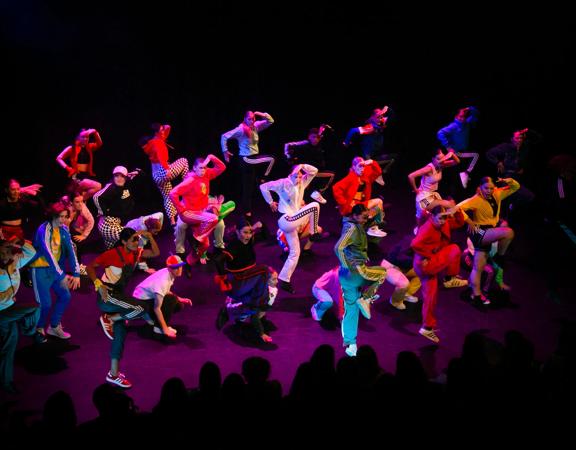 A group of 25 dancers wearing colourful athletic wear perform on stage.