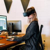 A person sits at a desk in an office using a Virtual Reality headset. 