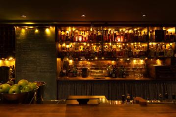 The bar at Dee's Place, a cocktail bar in Te Aro, Wellington. The dim space has amber lighting, many liquor bottles on the back shelf and a bowl of green apples on the counter.