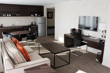 A modern hotel suite with a living room area, workspace, kitchenette and small dining space.