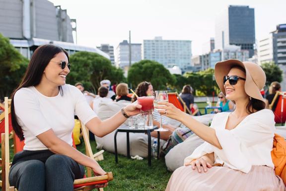 Two women sit on a grassy lawn, clinking glasses on a summers day with other people, trees and buildings in the background.