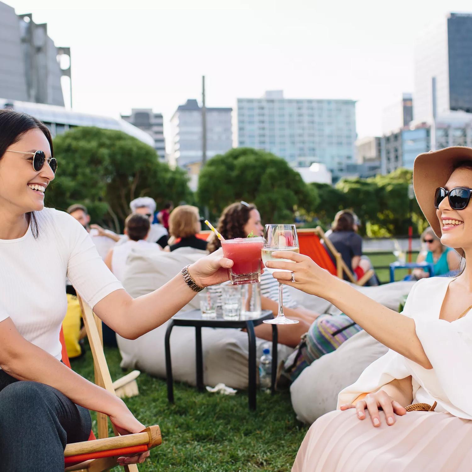 Two women sit on a grassy lawn, clinking glasses on a summers day with other people, trees and buildings in the background.